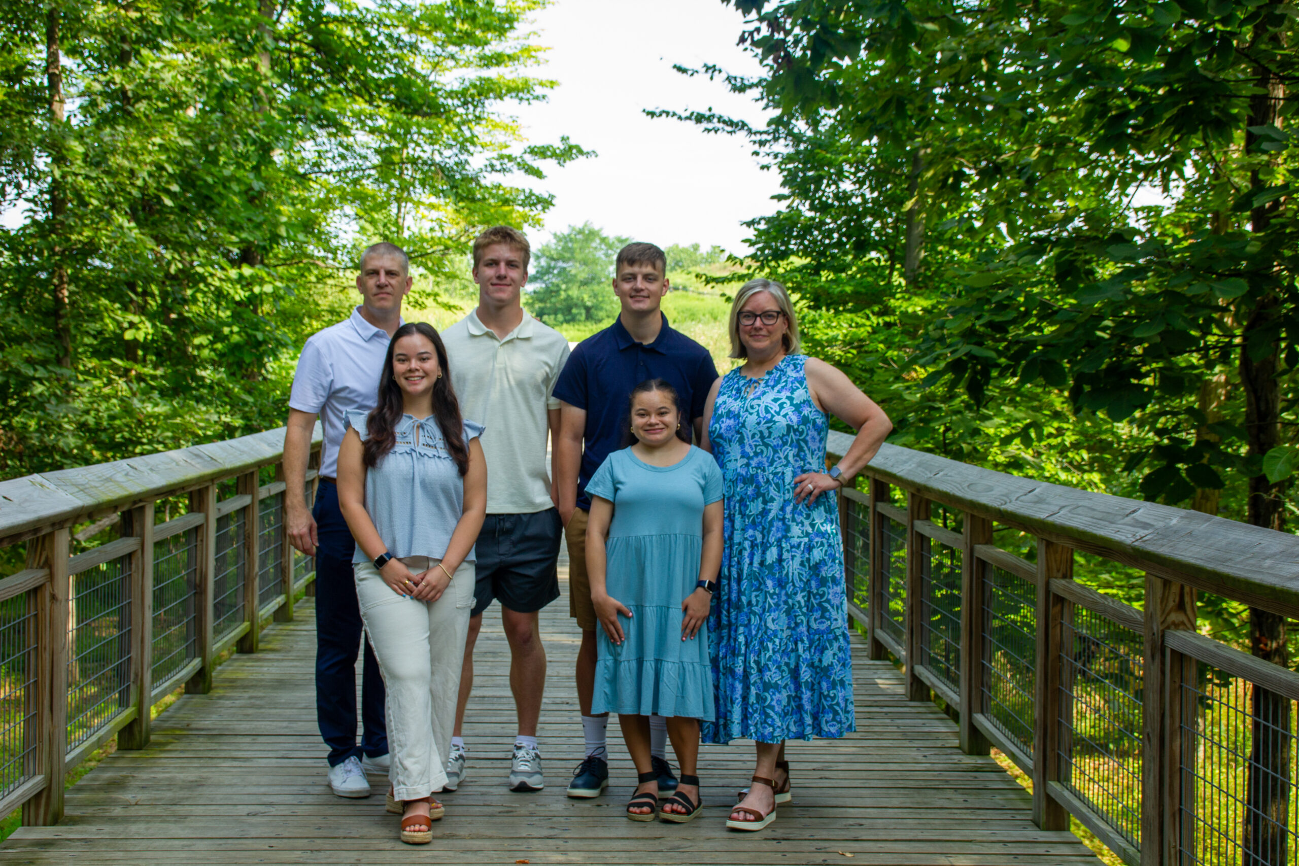 Family of six standing together on a wooden boardwalk surrounded by lush green trees on a bright day. Two adults and four children face the camera smiling, dressed in casual summer outfits, with sunlight filtering through the foliage behind them.