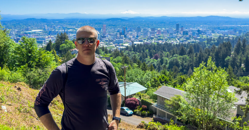 Man wearing sunglasses stands on a hillside overlook with a city skyline and a snow capped mountain in the distance on a clear sunny day. The elevated viewpoint shows dense green trees, residential rooftops, and an expansive urban landscape below.