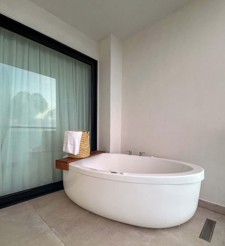 Freestanding bathtub beside a window at AVA Resort Cancun with a woven basket holding towels on a wooden ledge.