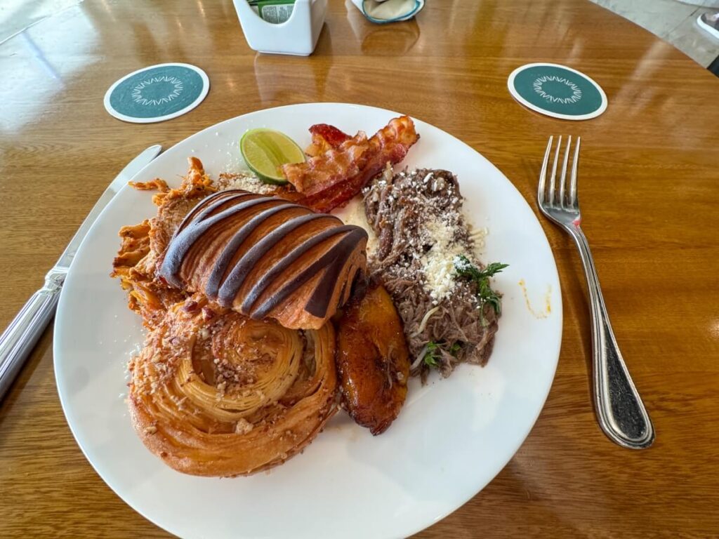 Breakfast plate at AVA Resort Cancun with pastries, sliced bread, bacon, refried beans topped with cheese, plantains, and a lime wedge on a wooden table.