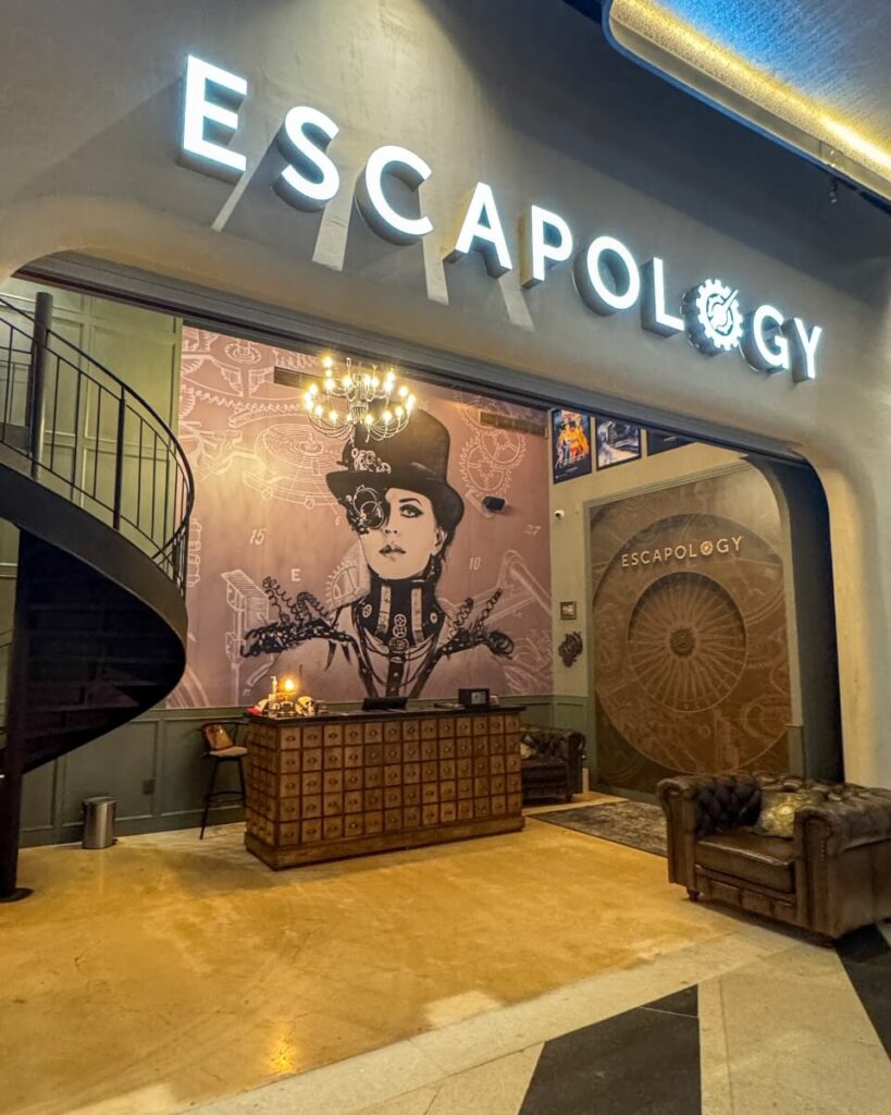 Entrance to "ESCAPOLOGY" at AVA Resort Cancun with a large mural of a woman in steampunk attire, a chandelier, and a reception desk below.