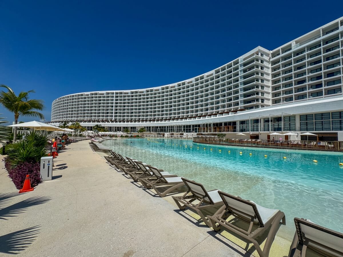 Large resort pool at AVA Resort Cancun lined with lounge chairs and a modern hotel building curving along the water.