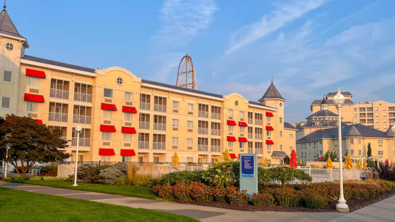 Hotel Breakers at Cedar Point with a cream colored exterior and bright red awnings lining the balconies under a blue sky. A tall roller coaster rises behind the hotel while landscaped paths and outdoor seating areas frame the waterfront resort setting.
