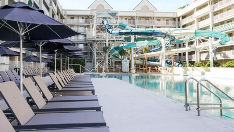 Pool area with lounge chairs and umbrellas beside a large water slide structure in a resort setting, showcasing family friendly features at Disney Good Neighbor Hotels.